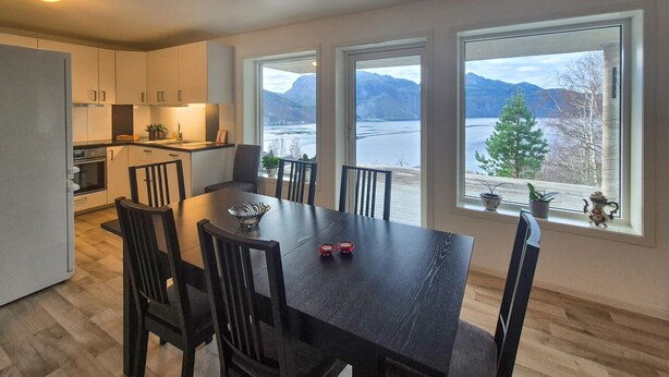 A modern kitchen with white cabinets, a dining area with a dark wooden table and chairs, and large windows offering a view of a lake and mountains.