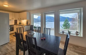 Une cuisine moderne avec des meubles blancs, un coin repas doté d’une table et de chaises en bois foncé, et de grandes fenêtres offrant une vue sur un lac et des montagnes.