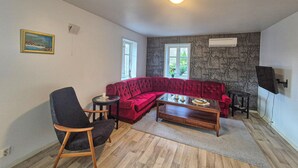 A living room with a red sofa, a wooden coffee table, a black armchair, and a small side table. There are two windows providing natural light, a wall-mounted TV, and a ceiling light.