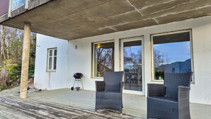 A covered outdoor area with two wicker chairs, a barbecue grill, and large windows offering views of the surrounding landscape.