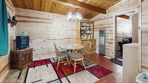 Een houten chalet van binnen, met een eettafel, stoelen, een kroonluchter, een klein kastje en een koelkast.