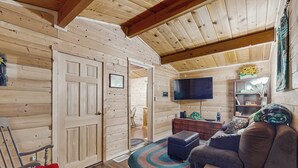 Een gezellig blokhutinterieur met een houten plafond, een aan de muur bevestigde televisie, een bank, een schommelstoel en een kleine tafel.