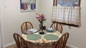 A dining area with a round table, wooden chairs, a floral tablecloth, a vase with flowers, a framed painting, and a window with a checkered curtain. - Comfortable Modern Stay Quiet 1BR Retreat Near Dayton, Eaton, Miami U& Richmond (Eaton)