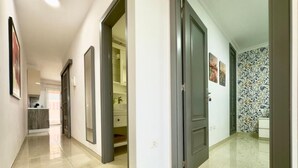 Um corredor com piso ladrilhado, uma porta que dá para um banheiro e uma cozinha visível através de uma porta aberta.