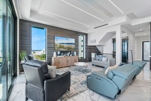 A modern living room with a large TV, leather sofas, a wooden cabinet, and a view of the outdoors. - Luxury Gulf-Front Sanctuary | Private Pool, Spa & Walk to Downtown (Fort Myers Beach)