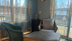 A room with a wooden floor, a grey sofa, a green chair, and a balcony with decorative curtains. - Lake View 2BR - 2-Min Walk to Sandy Beach - Sleeps 4-6 - Boyne City (Boyne City)