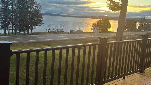 A wooden deck with a metal railing overlooking a lake, trees, and a distant view of a small building. - Lake View 2BR - 2-Min Walk to Sandy Beach - Sleeps 4-6 - Boyne City (Boyne City)