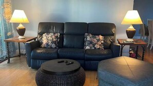 A modern living room with a black leather sofa, two side tables with lamps, a round wicker coffee table, and a blue ottoman. - Lake View 2BR - 2-Min Walk to Sandy Beach - Sleeps 4-6 - Boyne City (Boyne City)