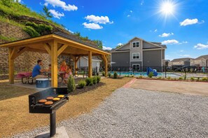 A covered outdoor area with a barbecue grill, picnic tables, and a swimming pool in the background. - Perfectly located for Branson fun, No step access, 2 pools and modern finishes. (Branson)
