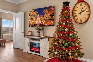 Une pièce avec un plancher en bois, une porte blanche, une cheminée, un sapin de Noël et une horloge au mur.