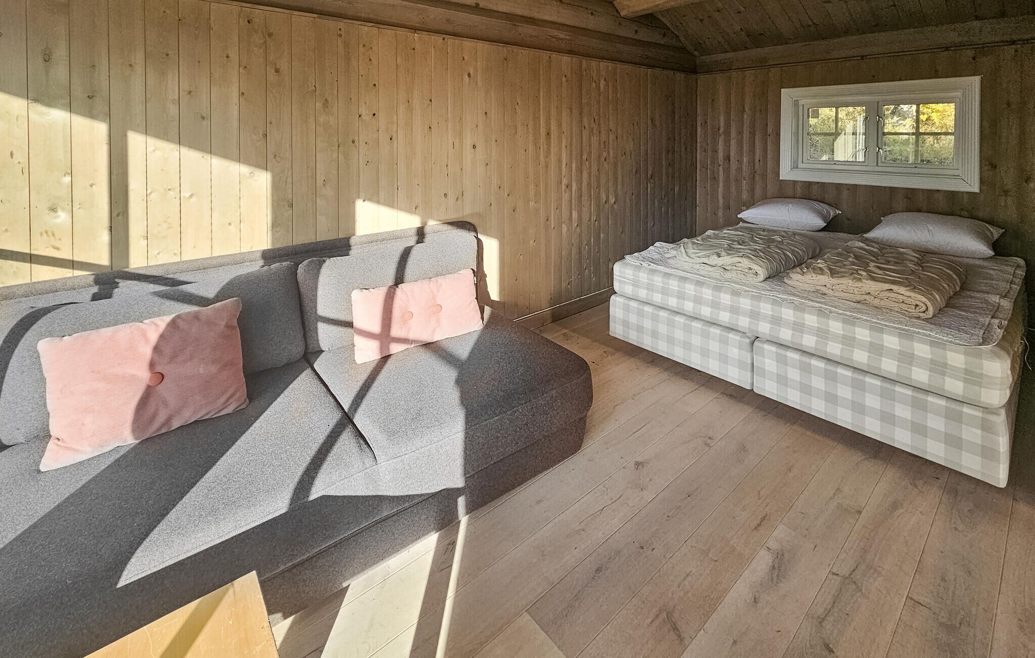 Ein gemütliches Zimmer mit einer grauen Couch, einem Bett mit karierter Bettdecke, Holzwänden und einem Fenster mit Blick auf Bäume.