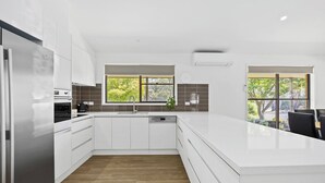 Une cuisine moderne avec des meubles blancs, un îlot central, un four encastré et un lave-vaisselle. Deux fenêtres laissent entrer la lumière naturelle et offrent une vue sur la verdure environnante.