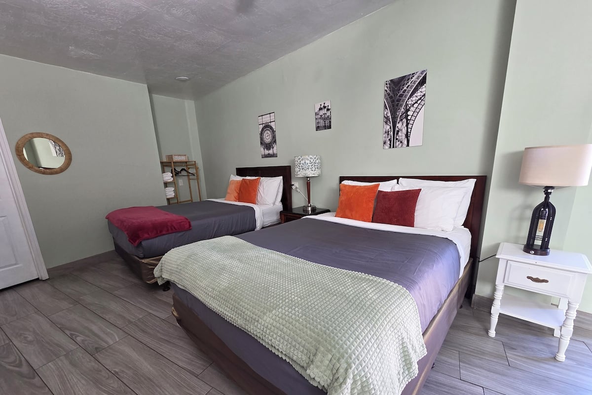A hotel room with two beds, a wooden floor, a mirror, a wardrobe, and a nightstand with a lamp.