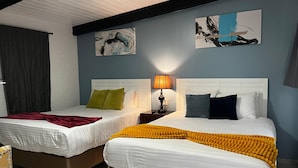 Ein Hotelzimmer mit zwei Betten, jeweils mit weißer Bettwäsche und Kissen in unterschiedlichen Farben. An der Wand hängen zwei Bilder, auf dem Nachttisch steht eine Lampe und es gibt ein Fenster mit Vorhang.