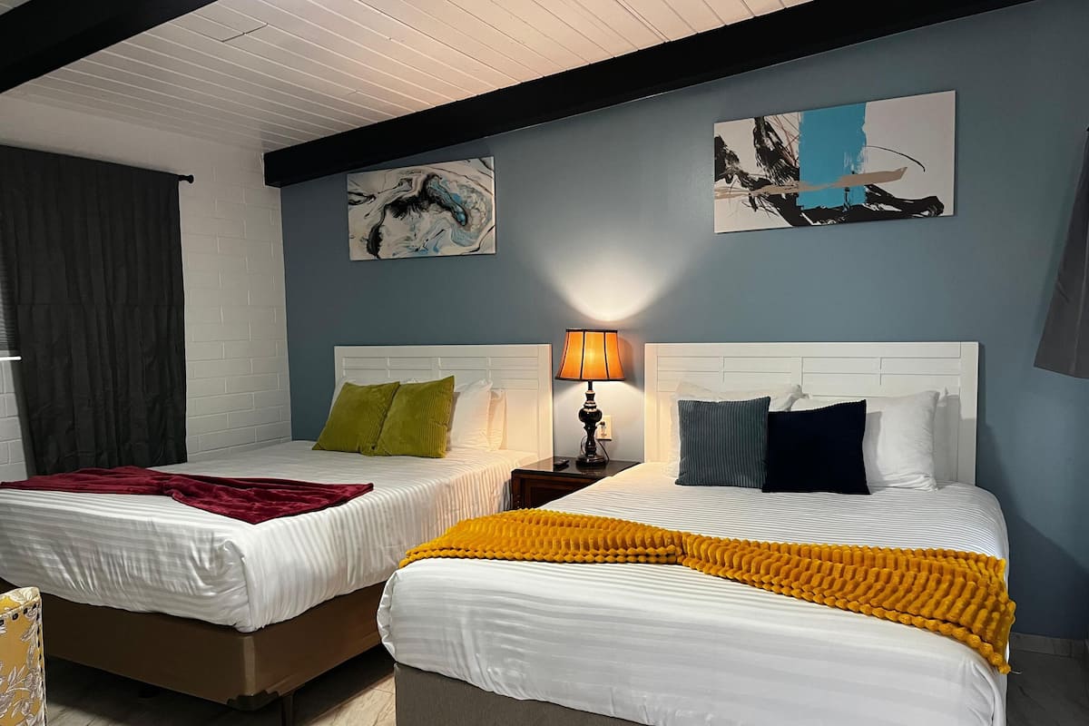 A hotel room with two beds, each with white bedding and different colored pillows. There are two paintings on the wall, a lamp on the nightstand, and a window with a curtain.