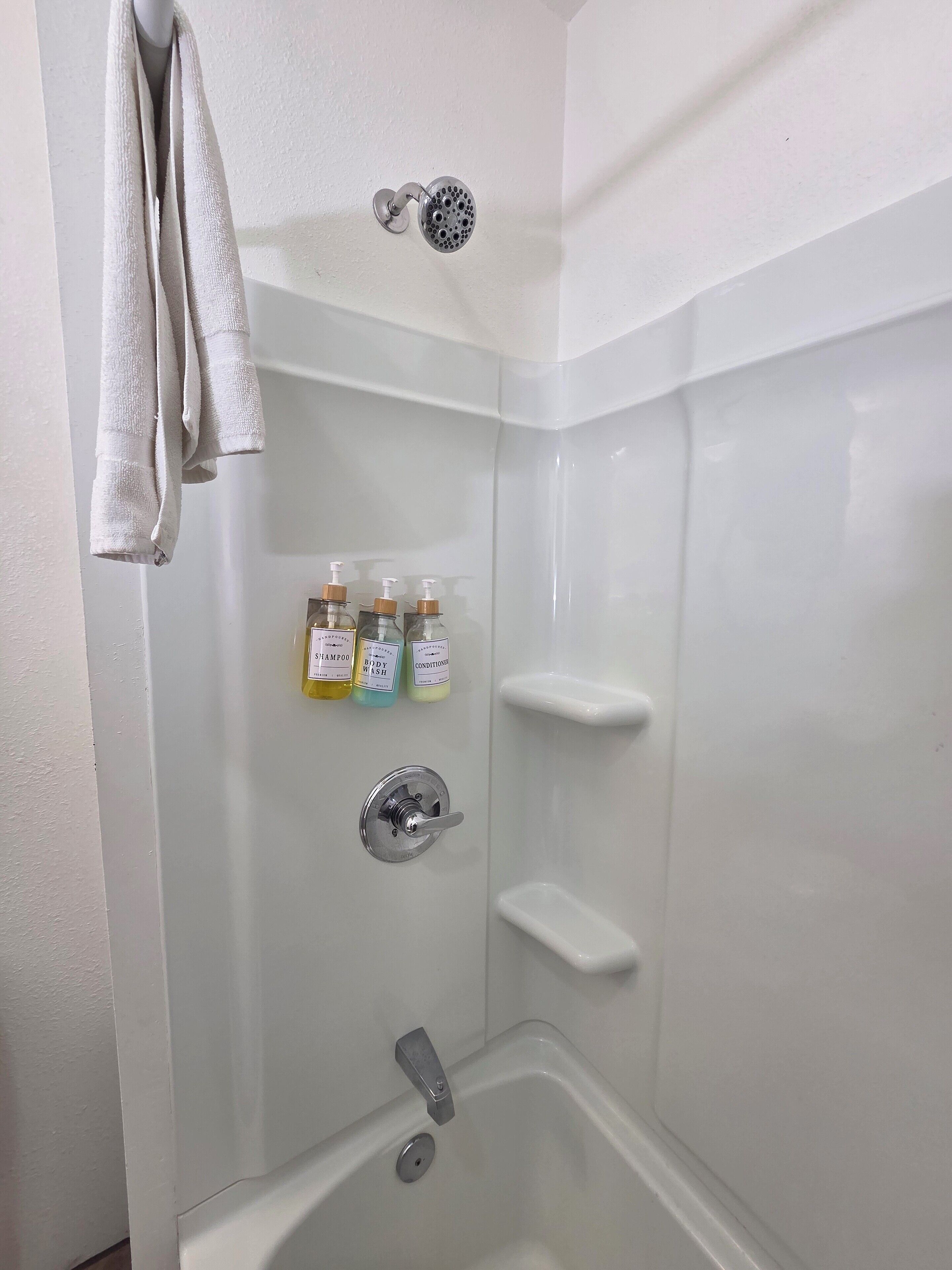 A bathroom with a shower, a towel rack, and three bottles of shampoo, body wash, and conditioner.