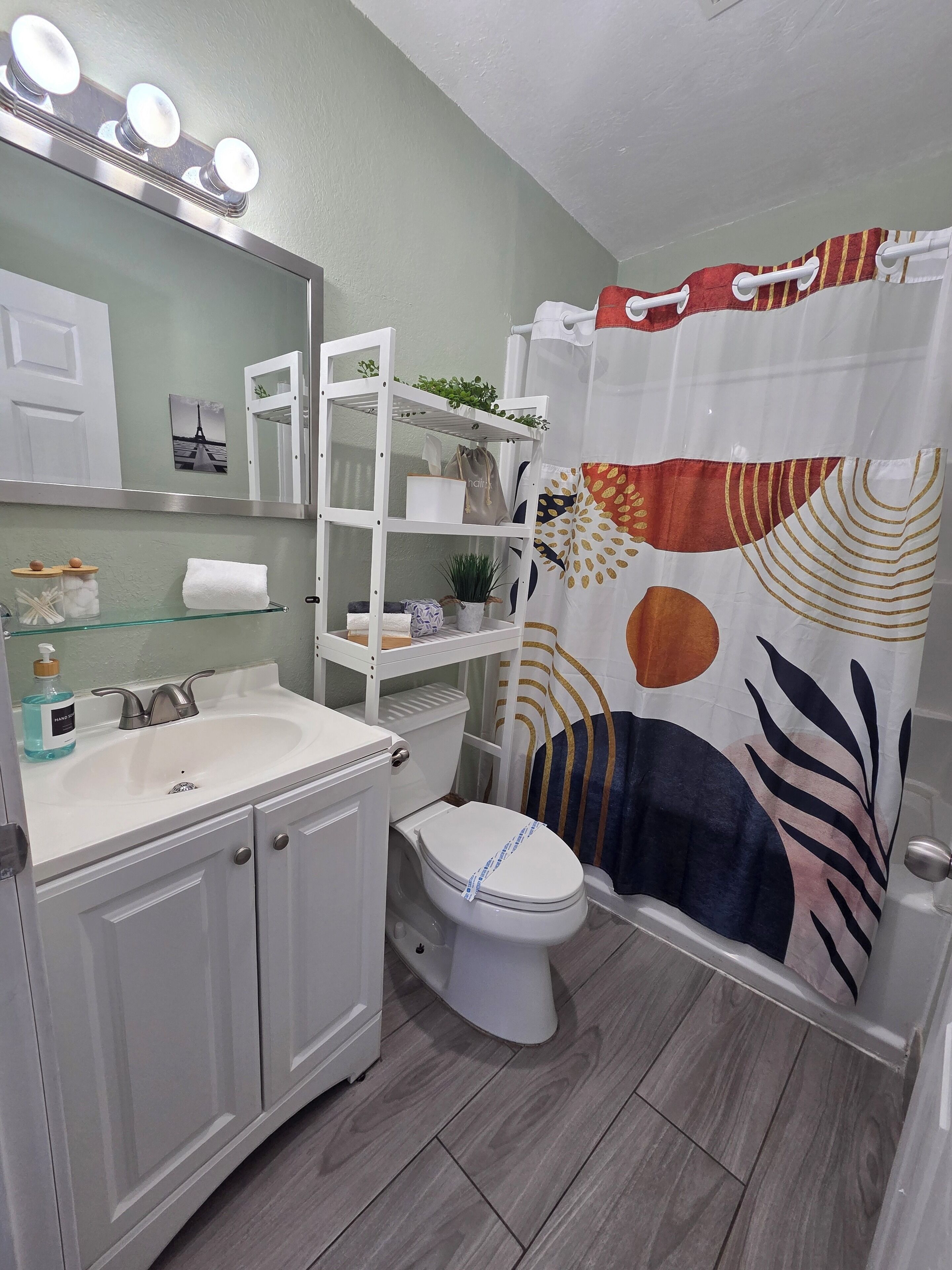 A bathroom with a white sink, mirror, and toilet. There is a shelf with toiletries and a shower curtain with a leaf pattern.
