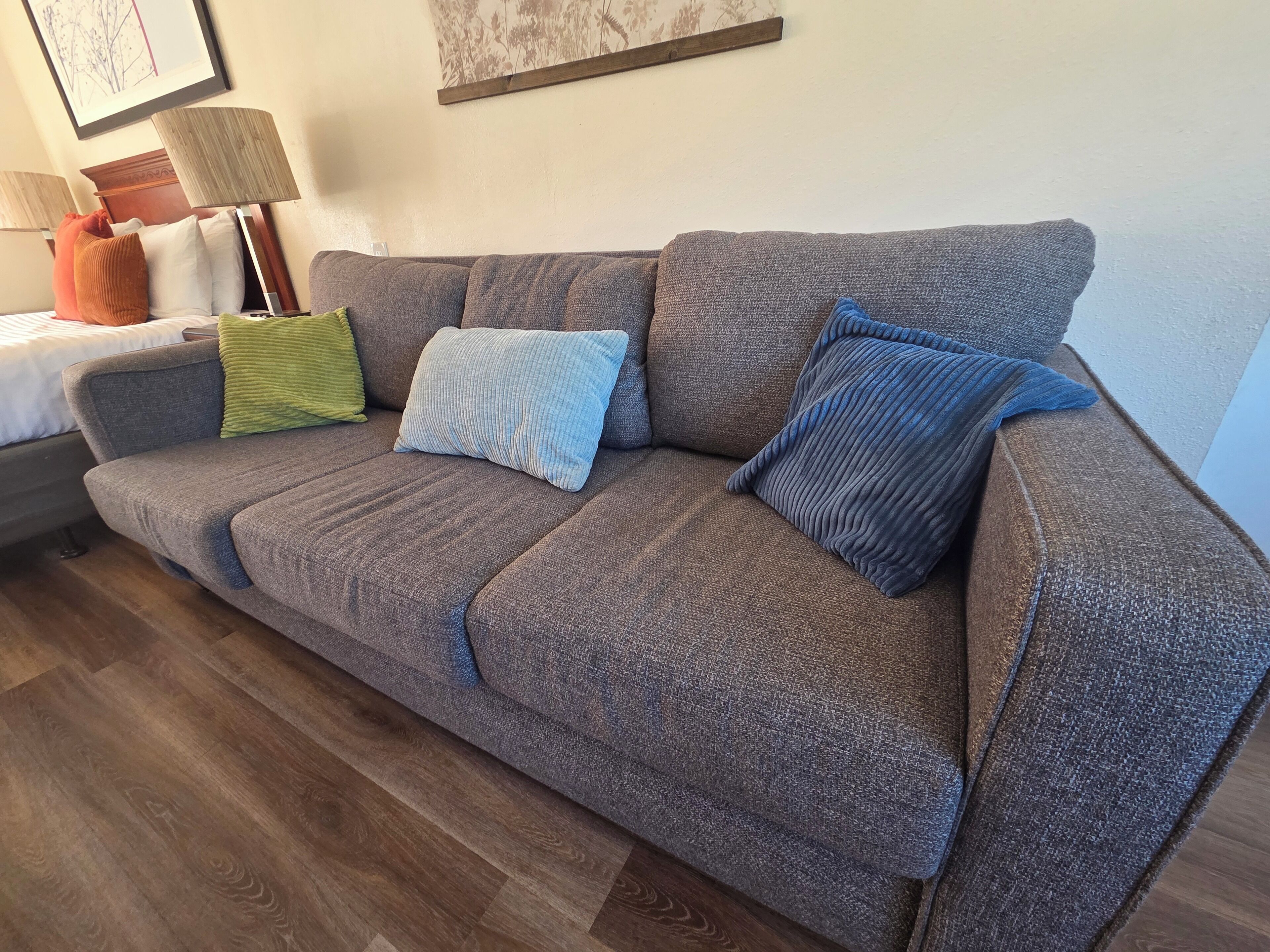 A grey sofa with three cushions in different colors, a wooden floor, and a lamp on a bedside table.