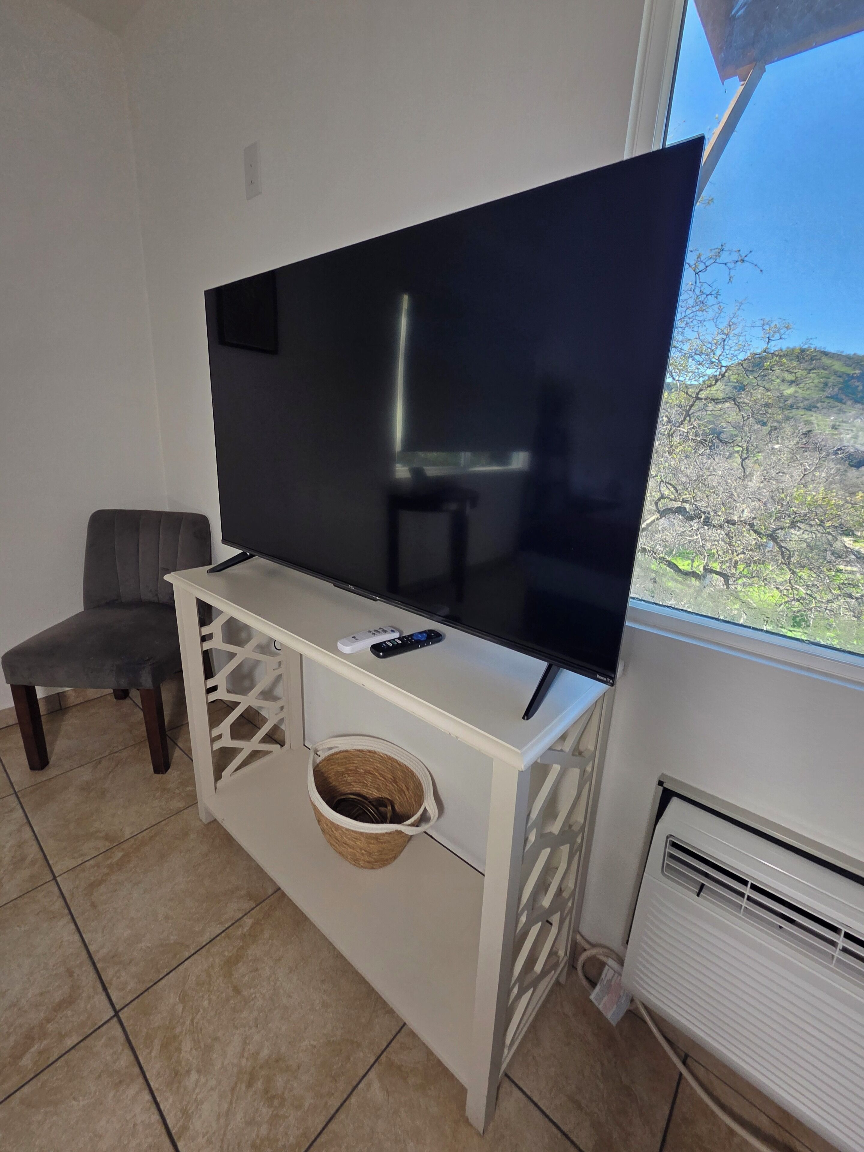 A living room with a flat-screen TV on a white cabinet, a grey chair, and a window with a view of trees.