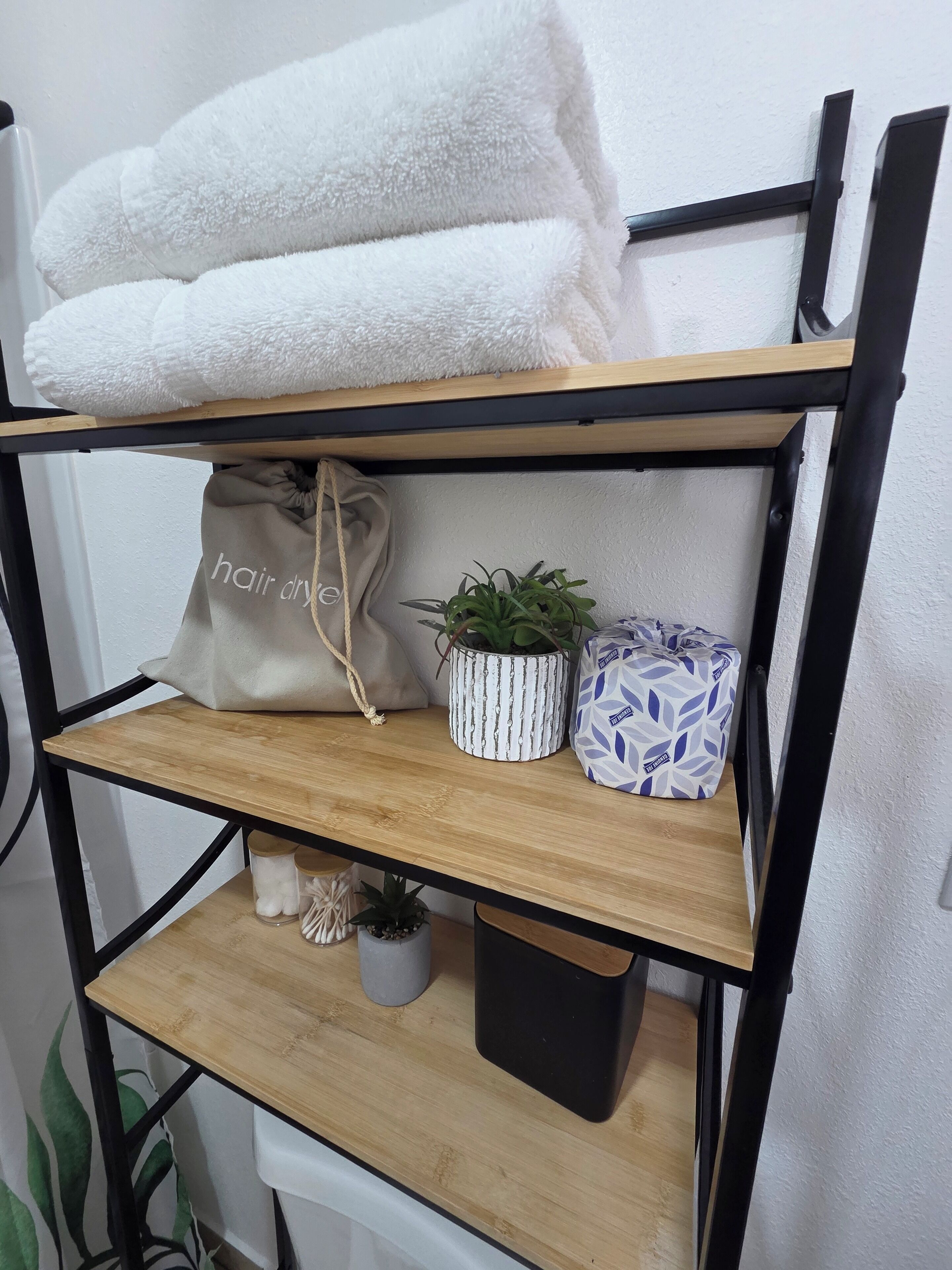 A shelf with a white towel, a hair dryer bag, a potted plant, and a patterned container.