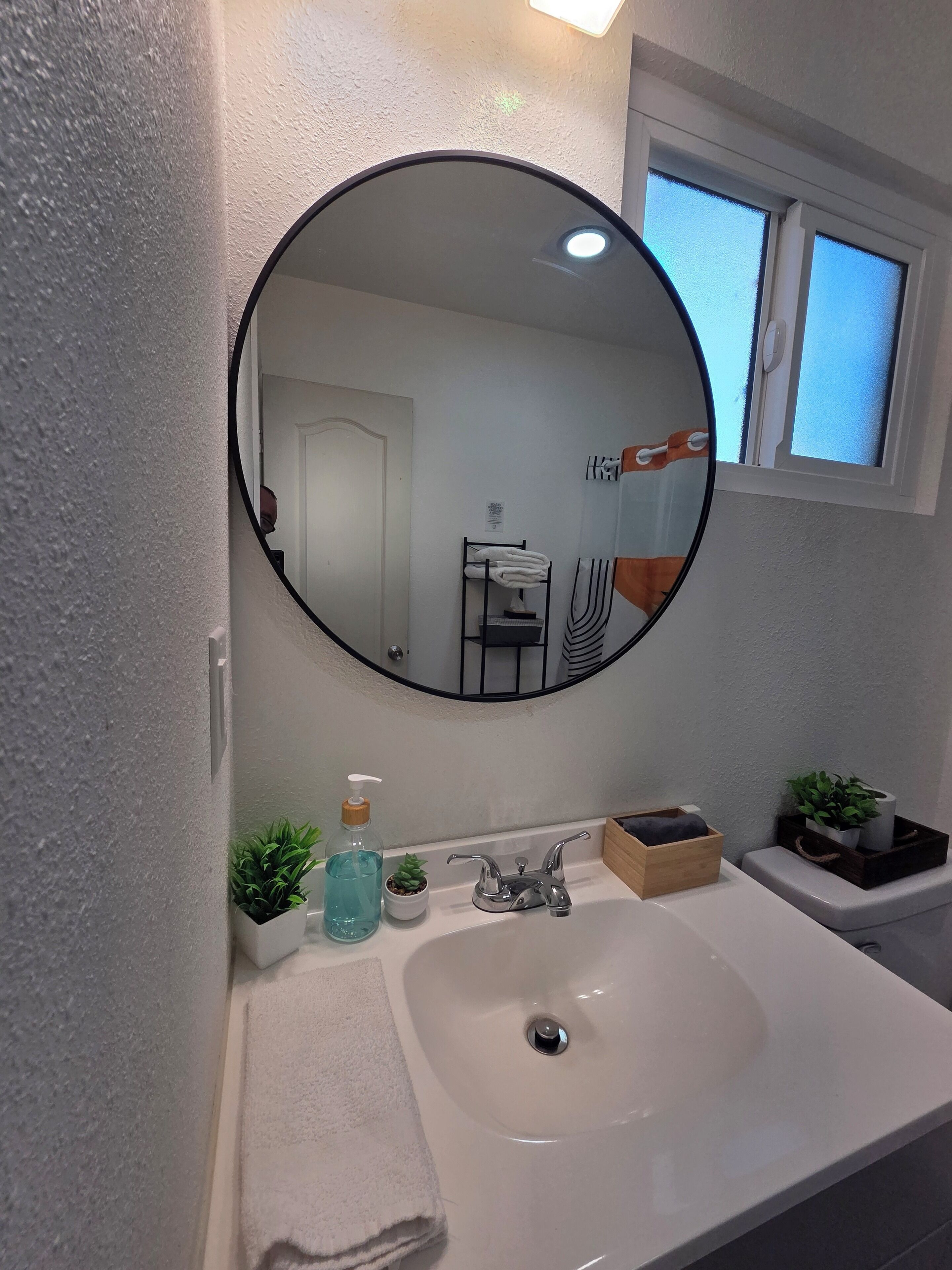 Un baño con un espejo redondo, un lavabo blanco, una toalla, un dispensador de jabón y una pequeña planta en maceta.