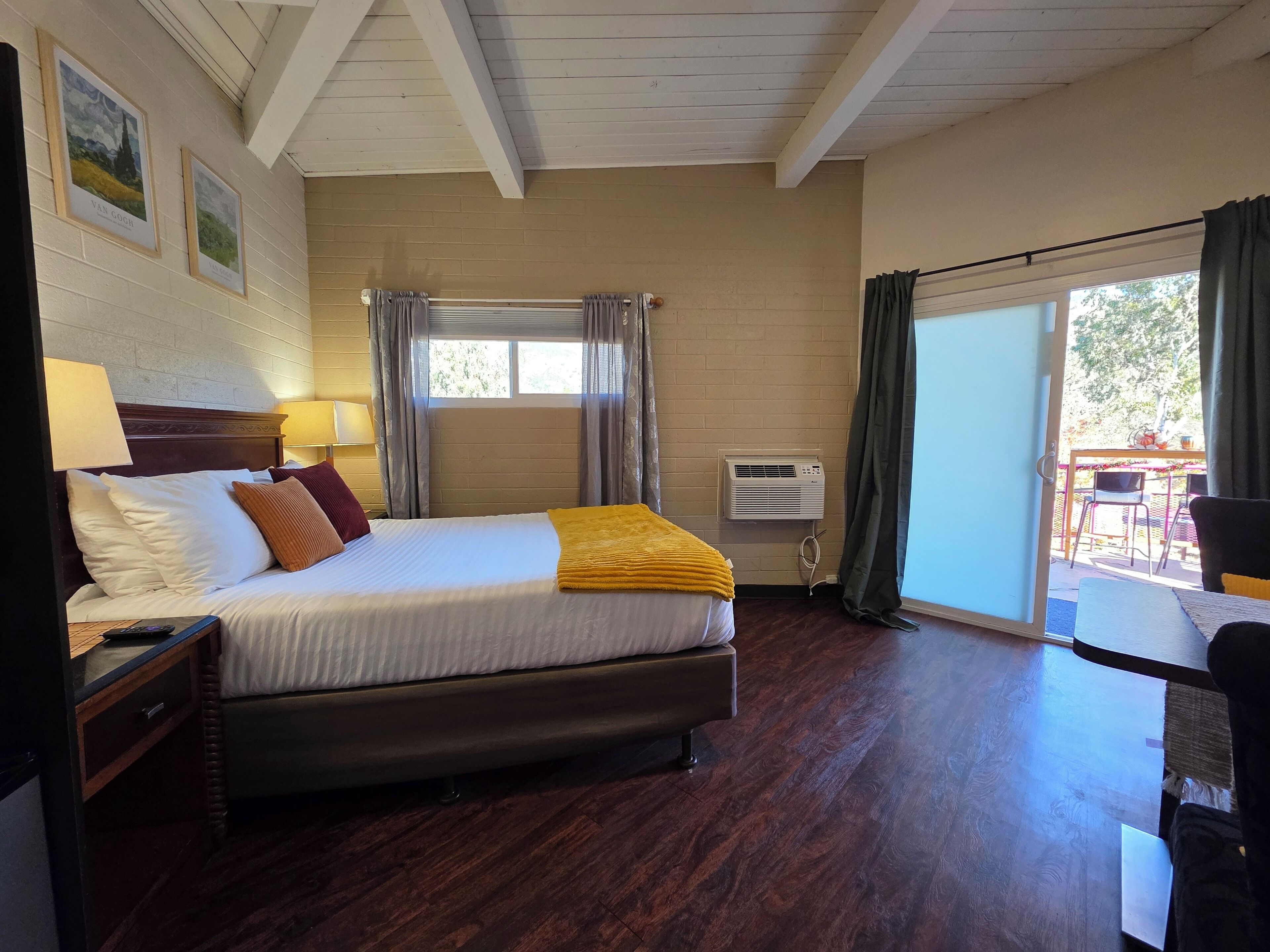 A hotel room with a bed, bedside table, lamp, air conditioning unit, and a view of the outdoors.