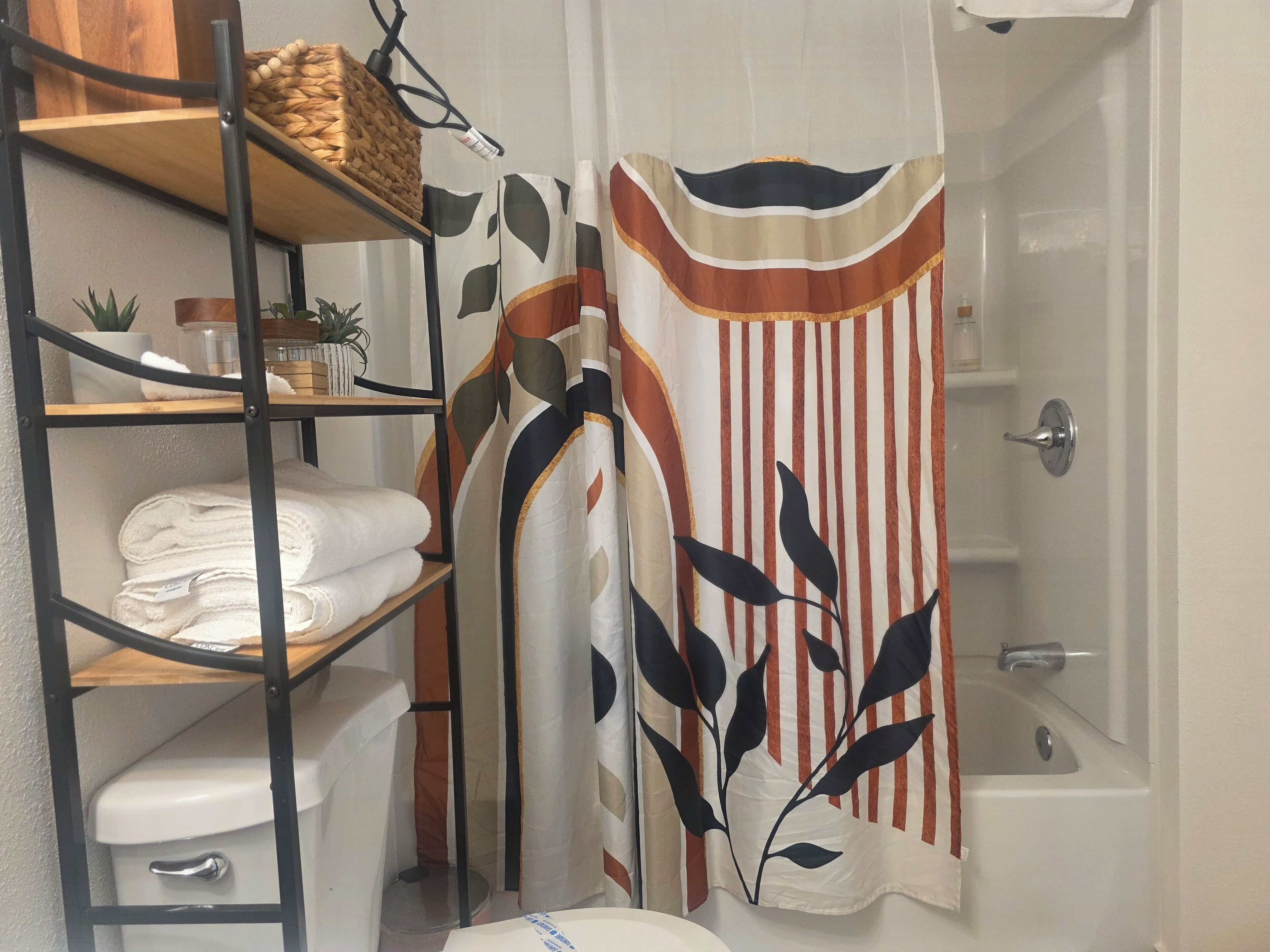 Een badkamer met douchegordijn met bladmotief, een toilet, een plank met handdoeken en een kleine potplant.