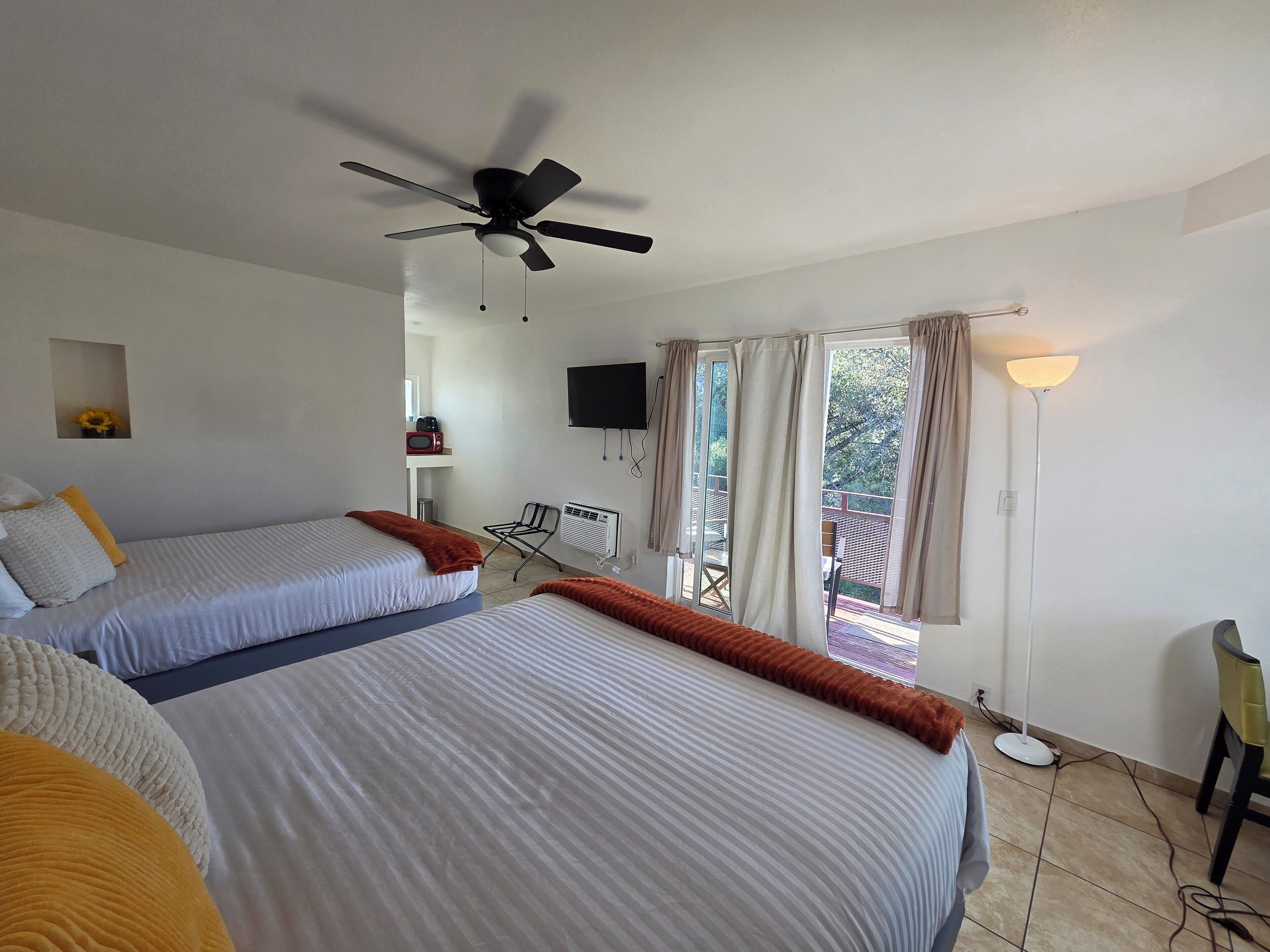 A hotel room with two beds, a ceiling fan, a TV, a window with curtains, a small table, and a chair.