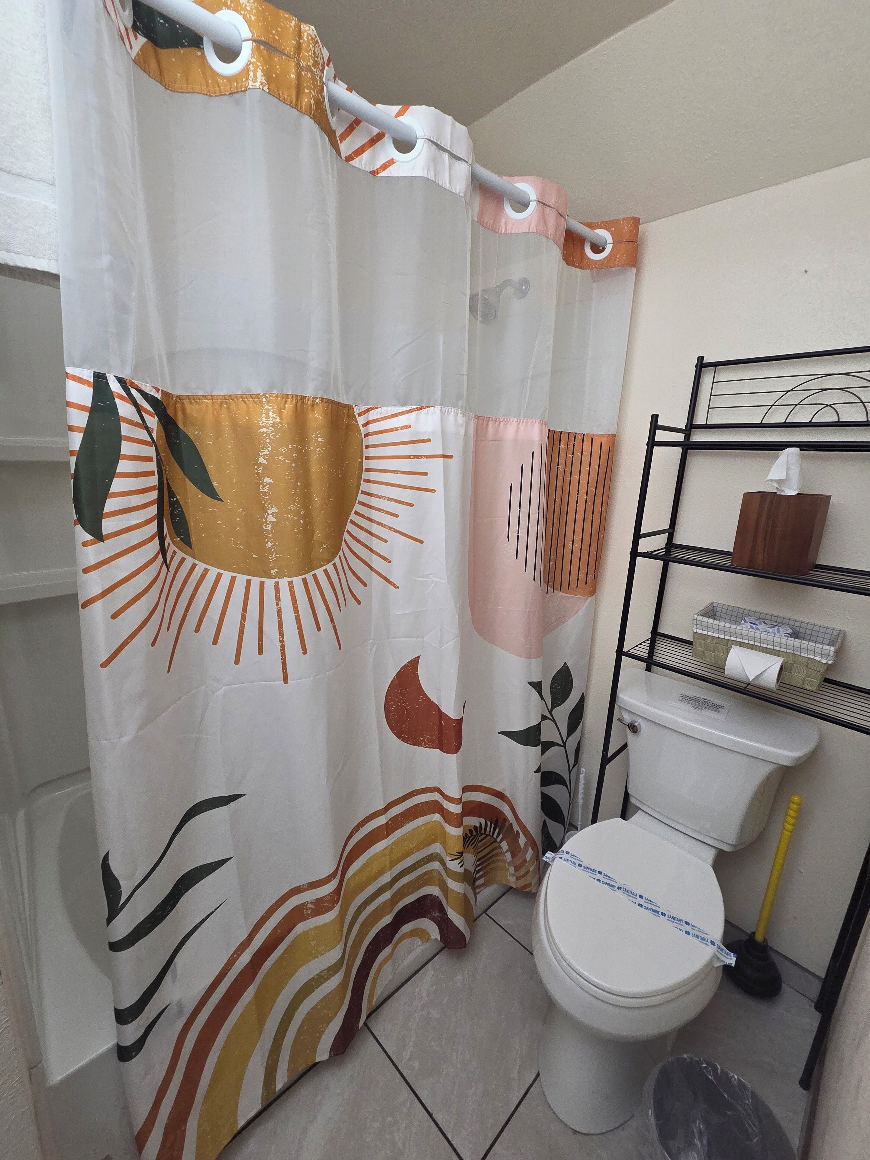 Un baño con inodoro, estante con artículos de tocador y cortina de ducha con diseño de sol y hojas.