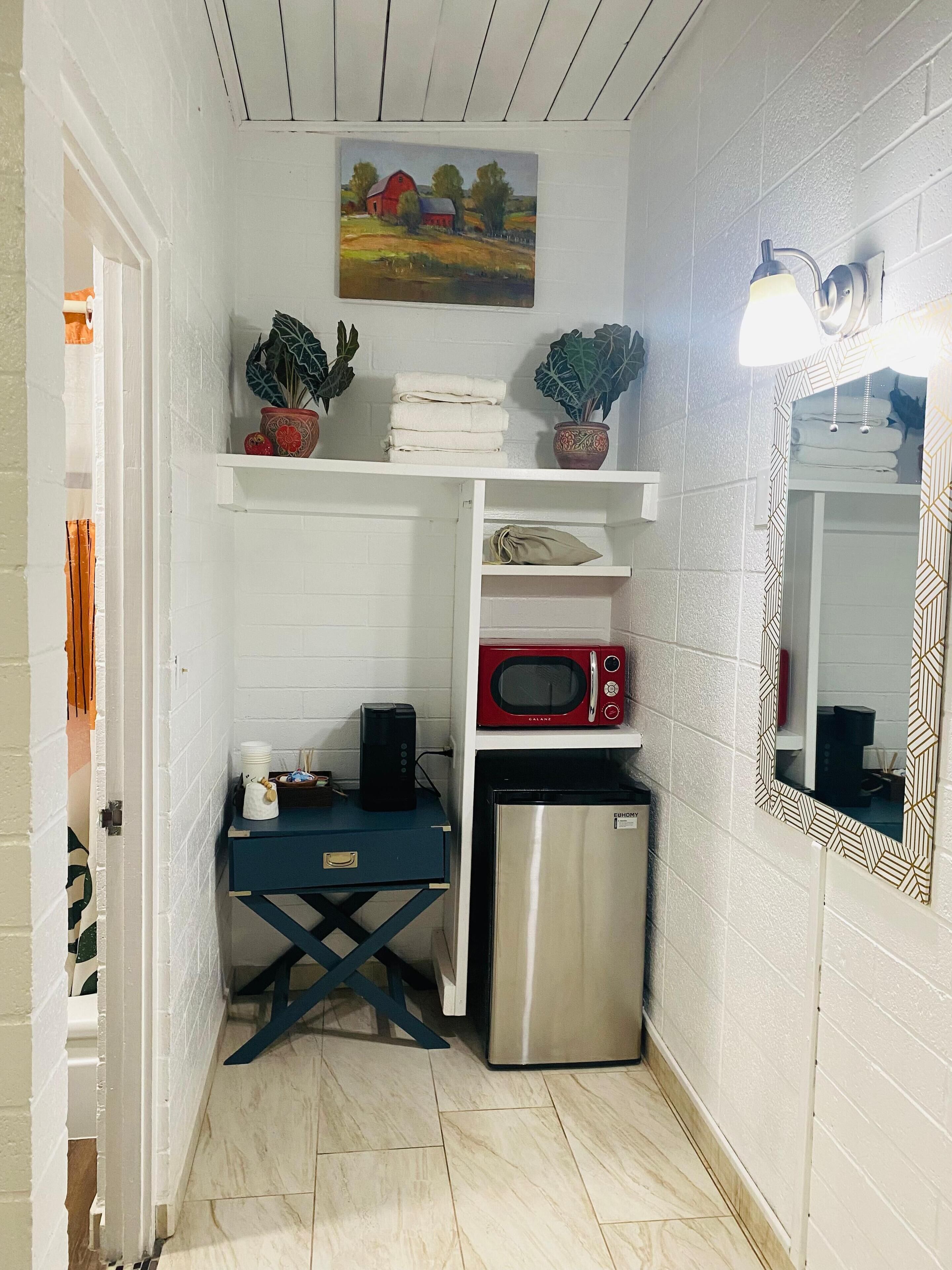 A compact kitchen with white walls, a painting of a rural scene, a microwave, a refrigerator, and a blue table with a kettle and plates.