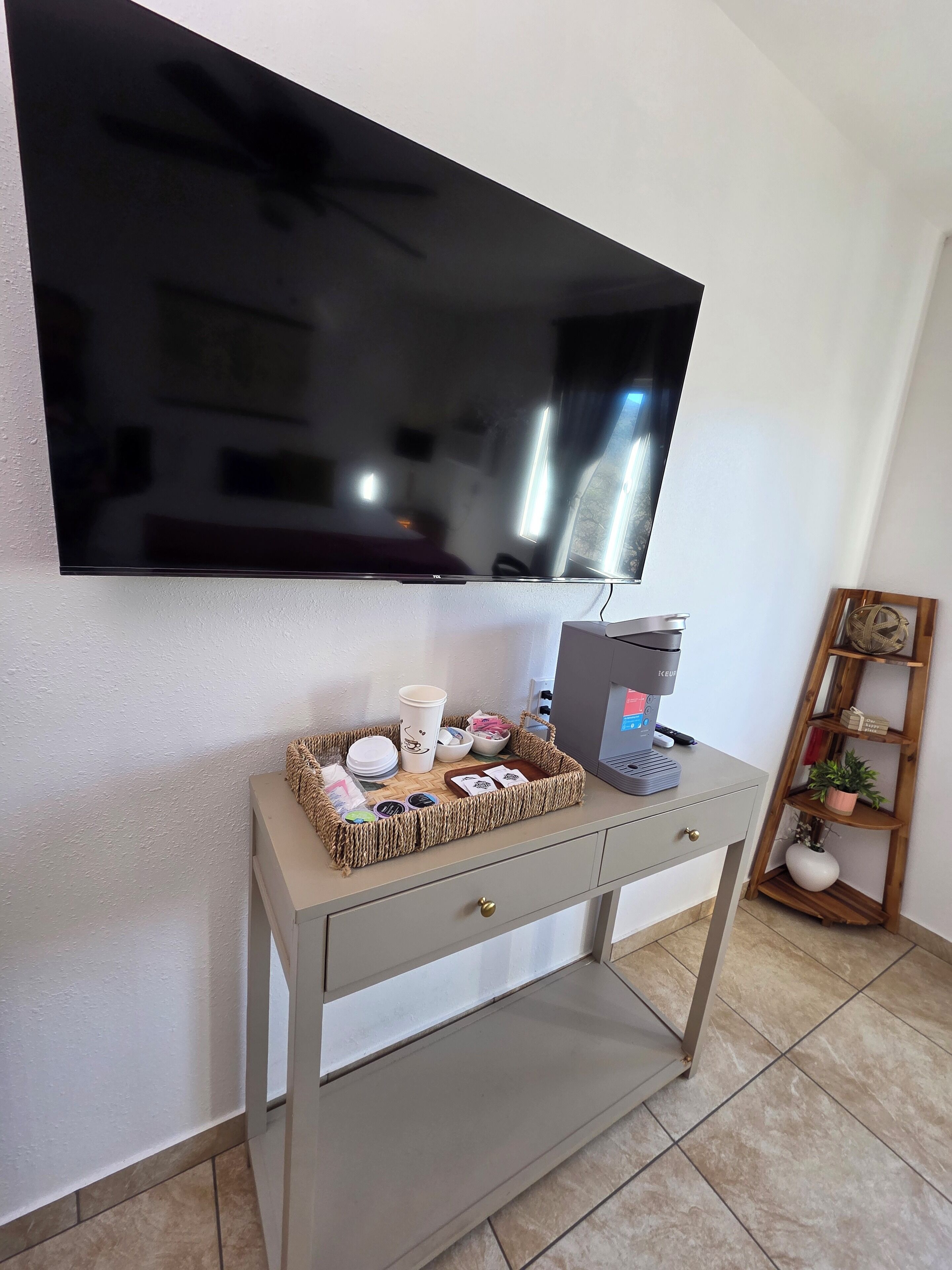 A modern living room with a flat-screen TV mounted on the wall, a coffee machine on a console table, and a small wooden shelf with decorative items.
