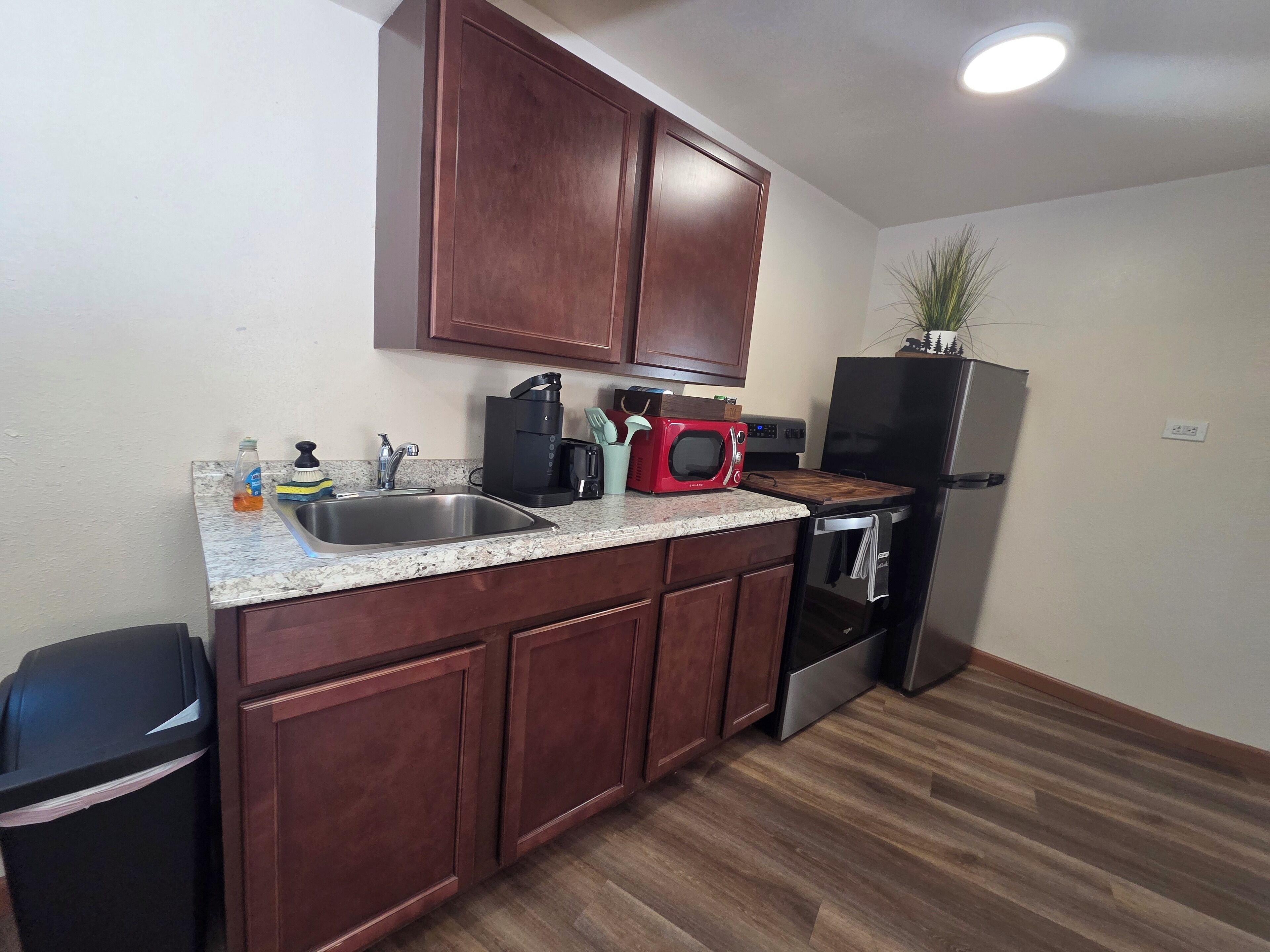 A compact kitchen with wooden cabinets, a stainless steel sink, a microwave, a toaster, and a refrigerator.