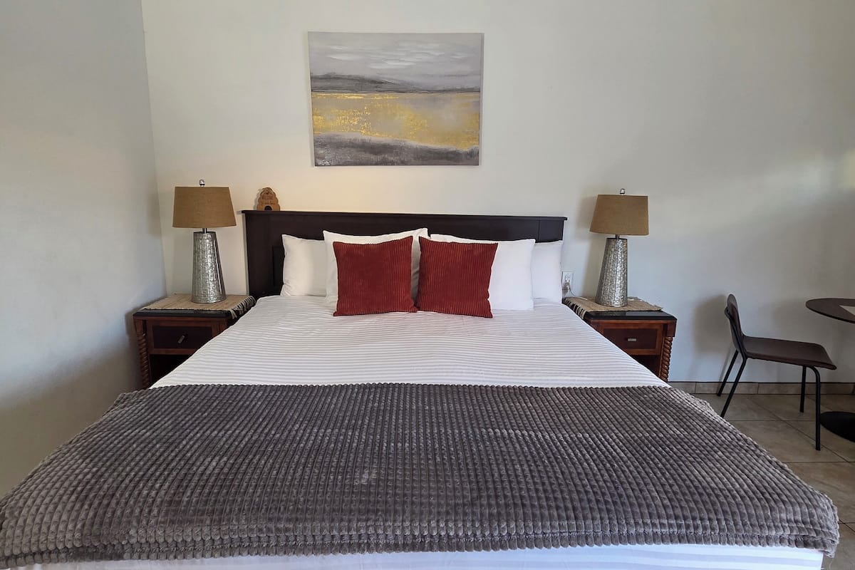 A neatly made bed with a grey blanket, two red pillows, and a white headboard. There are two bedside tables with lamps, a wall-mounted painting, and a ceiling fan.