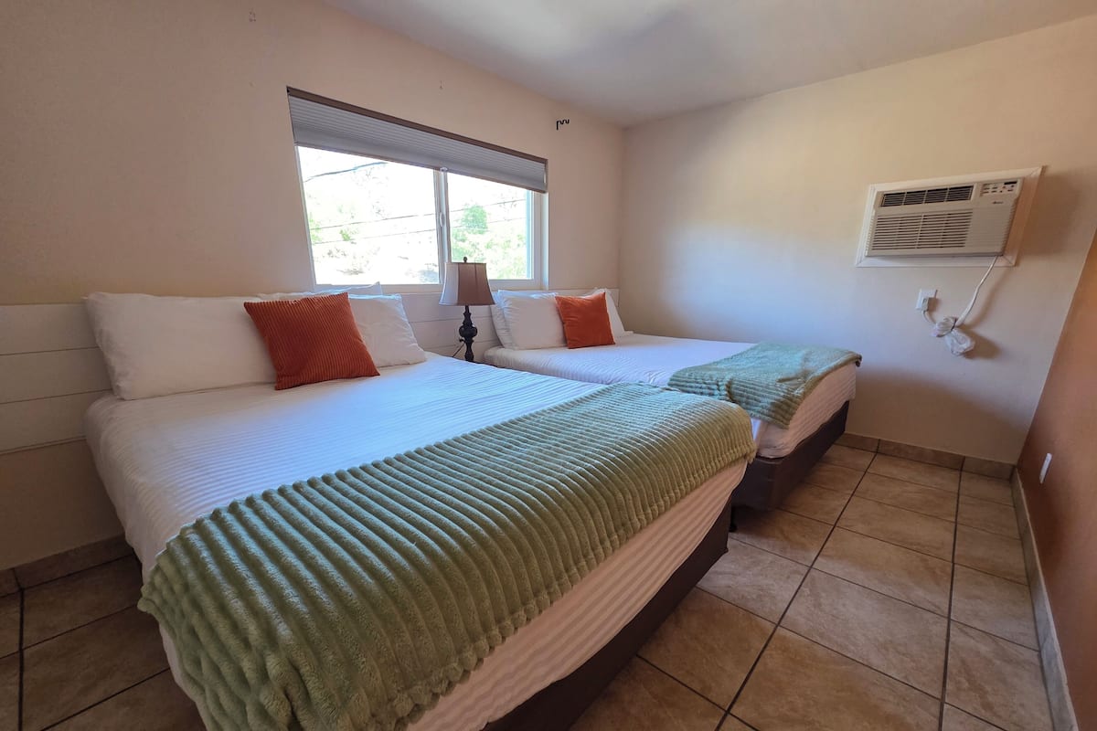 A hotel room with two beds, a window with blinds, an air conditioning unit, and a lamp.