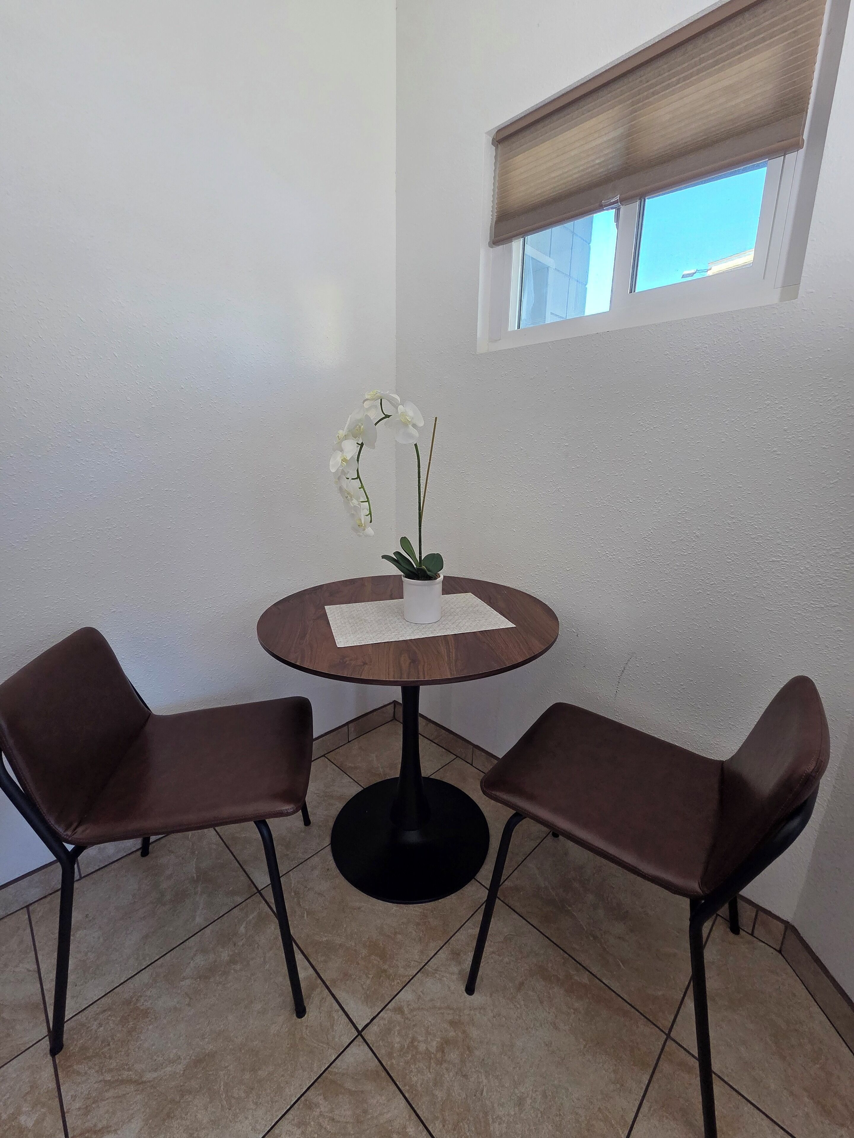 A small, round wooden table with two brown chairs, a potted orchid, and a window with a blind.