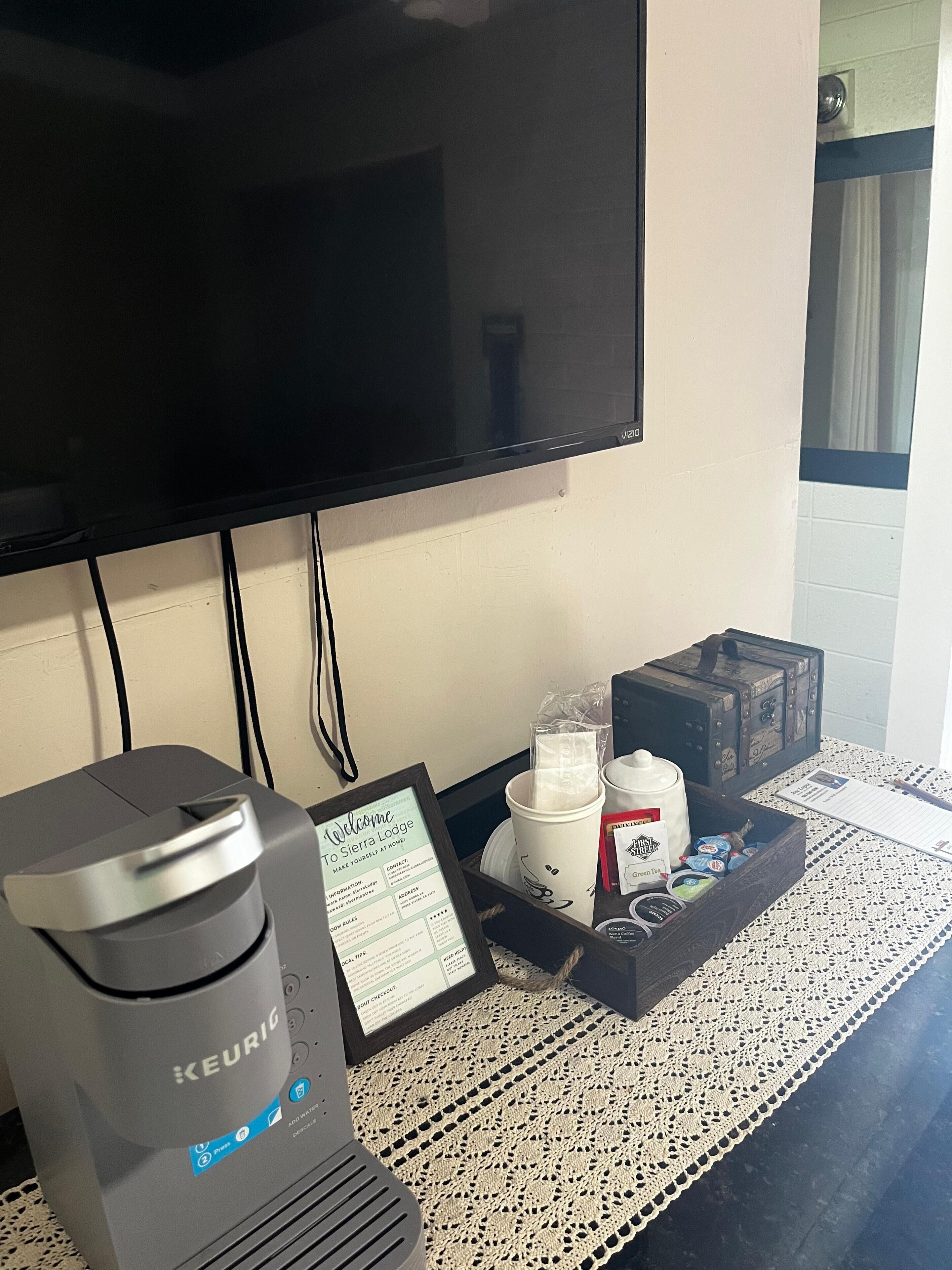 Una cafetera, un cartel de bienvenida, una bandeja con tazas y una caja de snacks, y un televisor en la pared.