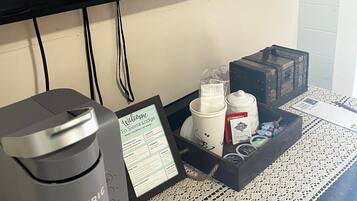 Una cafetera, un cartel de bienvenida, una bandeja con tazas y una caja de snacks, y un televisor en la pared.