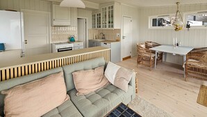 Un salon chaleureux avec un canapé gris, un coin repas avec des chaises en osier et une cuisine avec des meubles blancs et une bouilloire turquoise.
