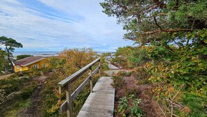 Un escalier en bois mène à une maison au toit rouge, entourée d’une végétation luxuriante et offrant une vue sur la mer.
