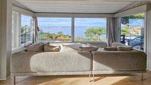 Et værelse med et stort vindue, der giver udsigt over vand og træer. Der er en sofa med puder og en gulvlampe.