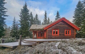 Une cabane en bois avec un toit de chaume, entourée d’une végétation recouverte de neige et de grands arbres.