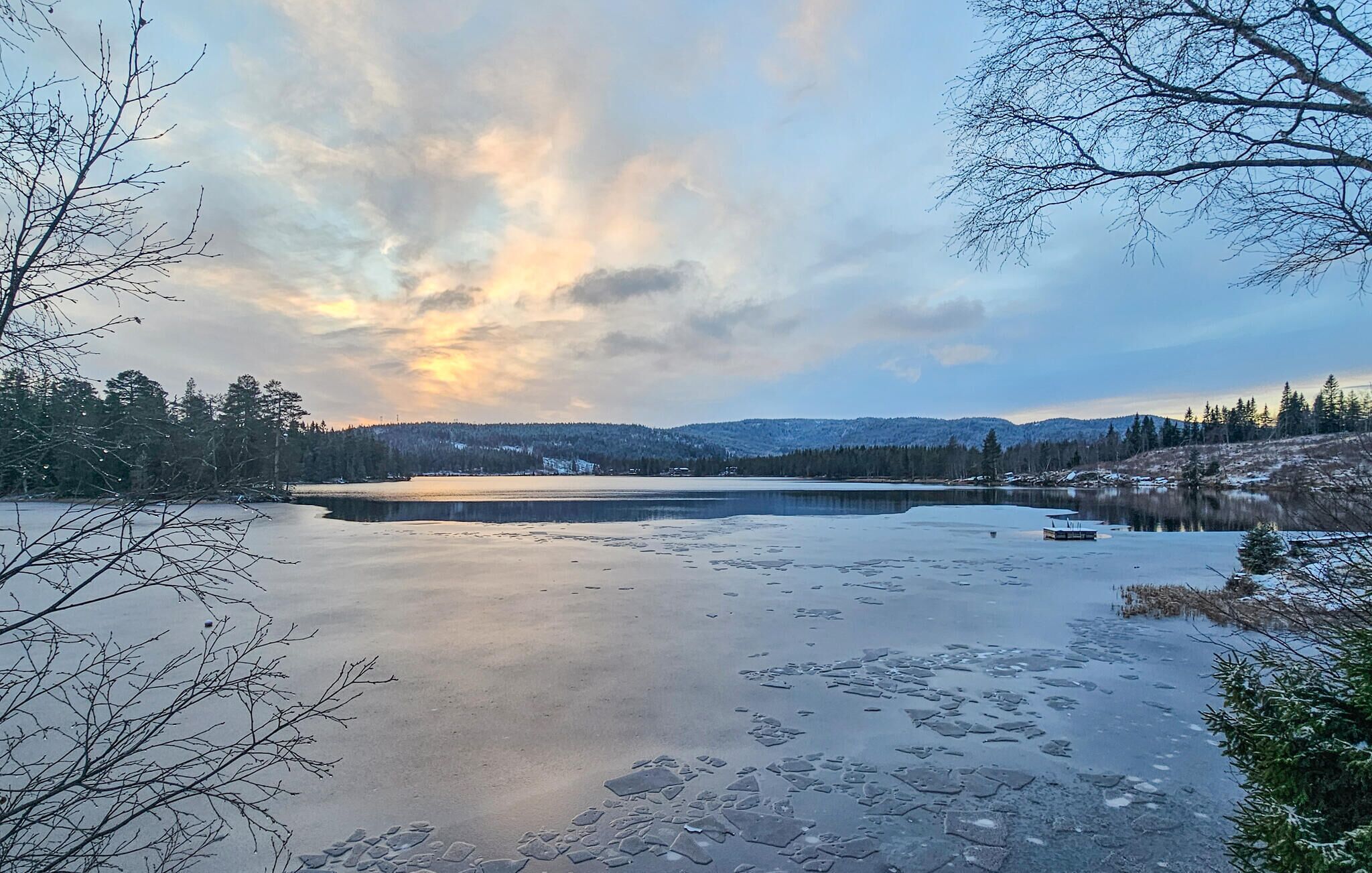 A frozen lake with a wooden dock, surrounded by trees and mountains in the background.