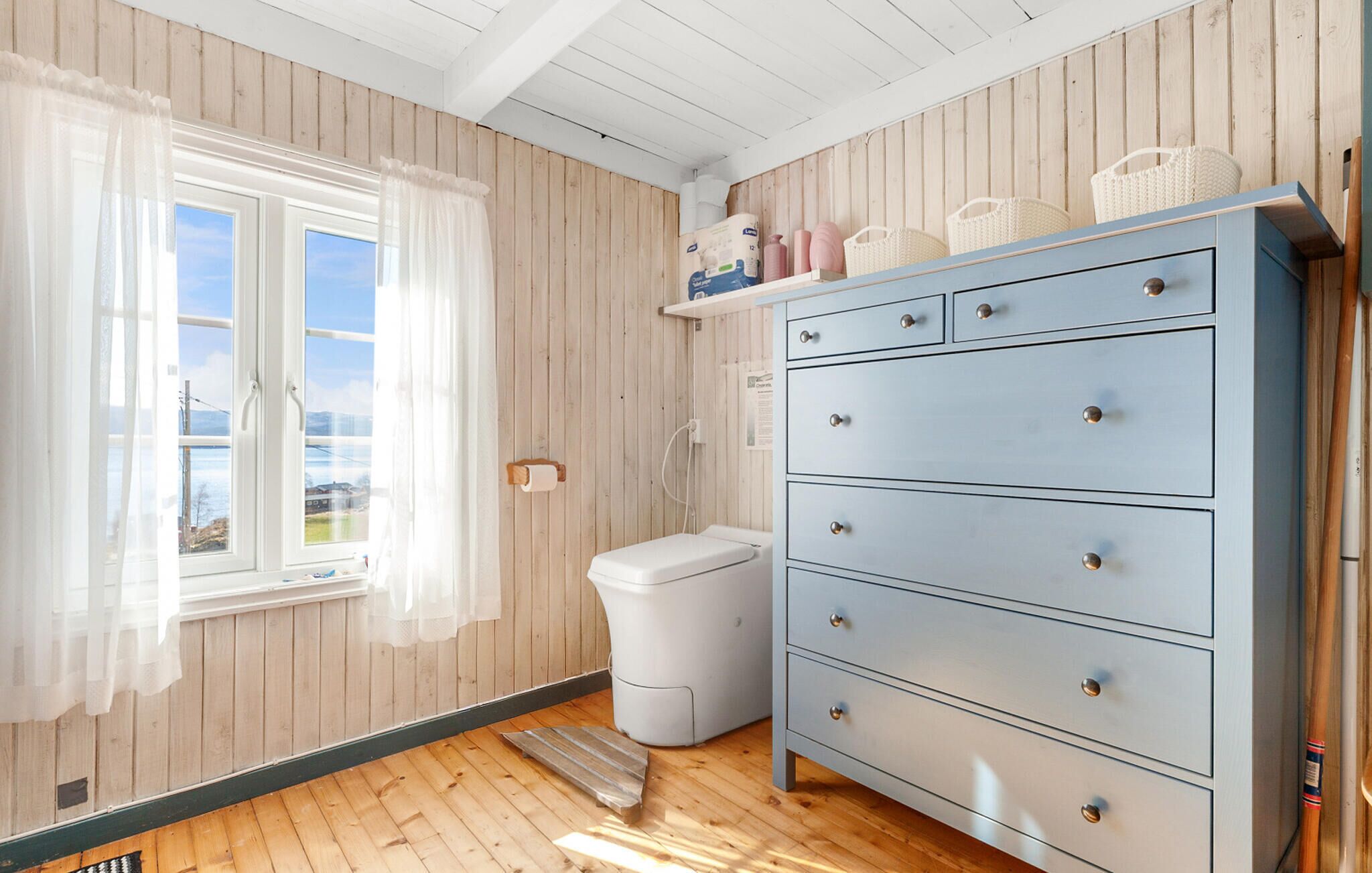 A room with wooden walls, a large blue dresser with drawers, a toilet, and a window with sheer curtains.