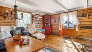 A cozy cabin interior with a wooden floor, a sofa, a chair, a dining table with a bowl and glasses, and a window with curtains.