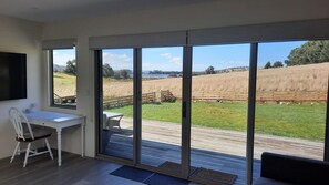 Une pièce comprenant un bureau, une chaise et de grandes portes-fenêtres coulissantes donnant sur une terrasse avec vue sur un champ verdoyant et une étendue d’eau.
