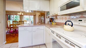 Una cocina con armarios blancos, una estufa y un hervidor. Hay una zona de comedor con sillas y una mesa.