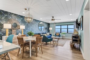 A modern living room with a dining area, featuring a large window, ceiling fan, and decorative wall art. - Relaxing Oceanfront Condo w/ Pool, Balcony & Sunrise Views (New Smyrna Beach)