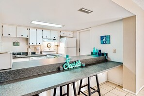 A modern kitchen with white cabinets, a granite countertop, and a 'Seaside' sign. - Oceanfront Condo with Pool & Patio – Peaceful Beachside Vibes (New Smyrna Beach)