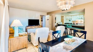A modern living room with a TV, sofa, and dining area with chairs and a table. - Oceanfront Condo with Pool & Patio – Peaceful Beachside Vibes (New Smyrna Beach)