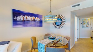 Uma sala de jantar com uma mesa de vidro, cadeiras e toalha de mesa estampada com flores azuis. Há um quadro com cisnes na parede e um lustre decorativo sobre a mesa.
